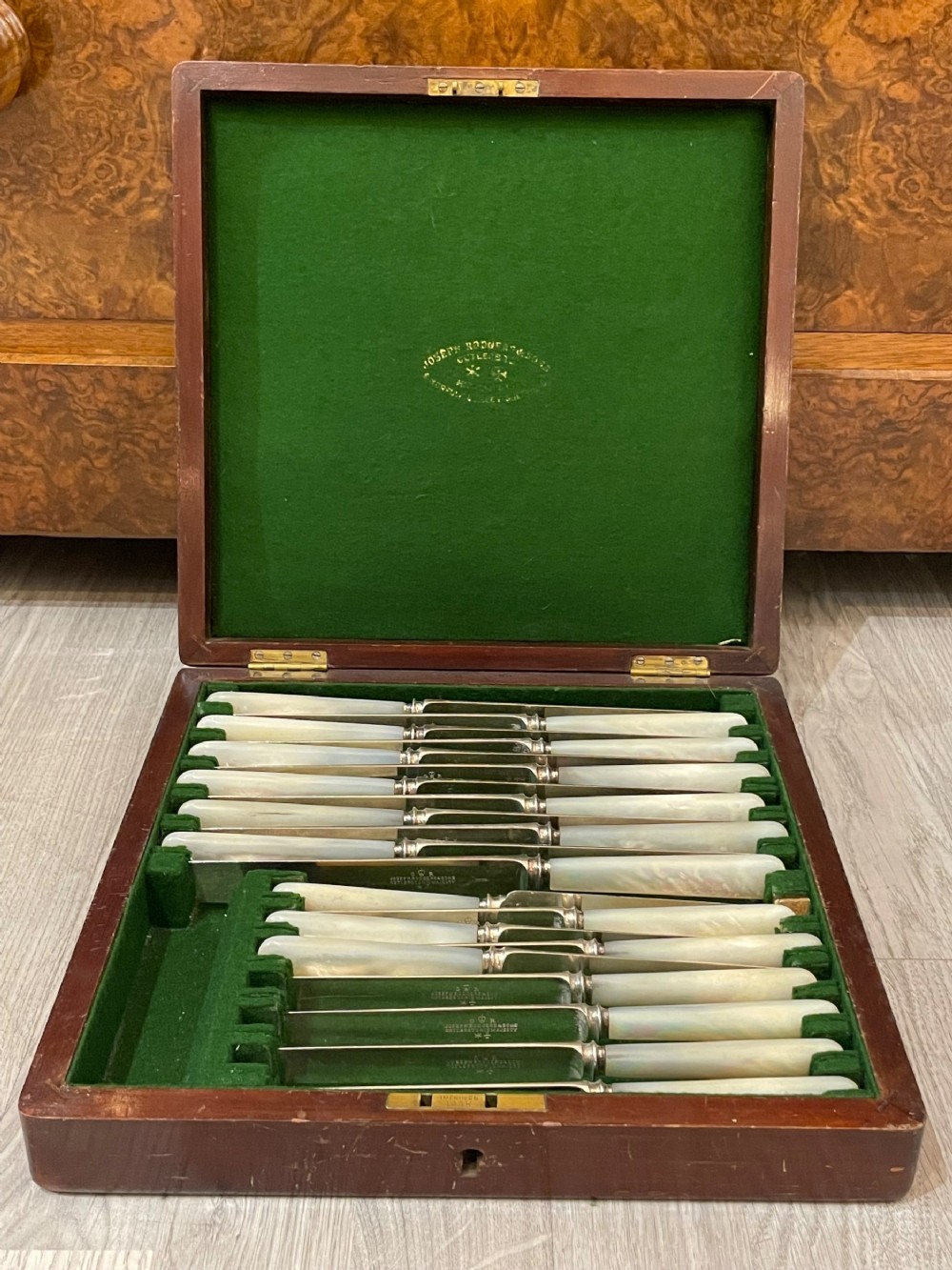 An open fitted mahogany case lined with green velvet holding steel knives with iridescent mother of pearl handles.
