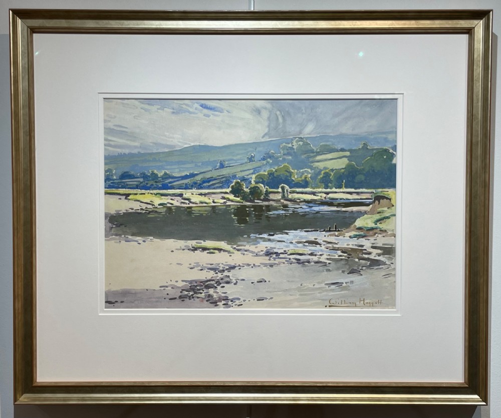 A framed watercolour painting depicting a river and rolling green hills under a cloudy sky, housed in a cream mount and metallic gold frame.