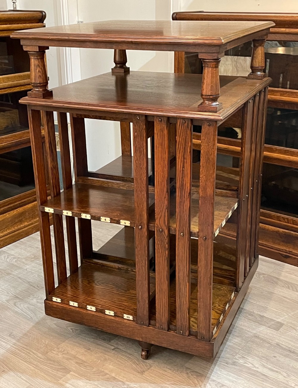 A multi-tiered Edwardian dark oak revolving bookcase with vertical side slats, upper gallery spindles, and small numbered tags along the shelf edges.