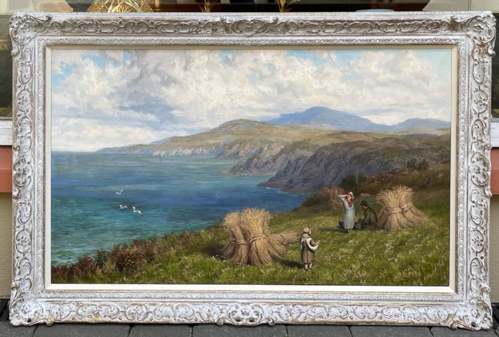 A large Victorian oil painting depicting a coastal landscape with figures resting near haystacks on a green cliff overlooking a blue ocean, framed in a carved swept frame.