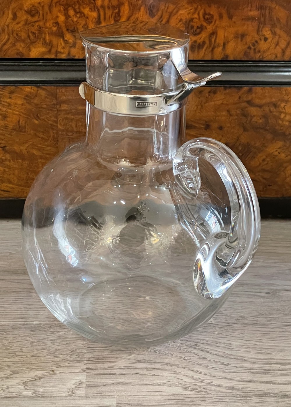 An Edwardian clear glass claret jug featuring a bulbous body, an integrated glass handle, and a polished silver-plated collar with a hinged spout lid. View II