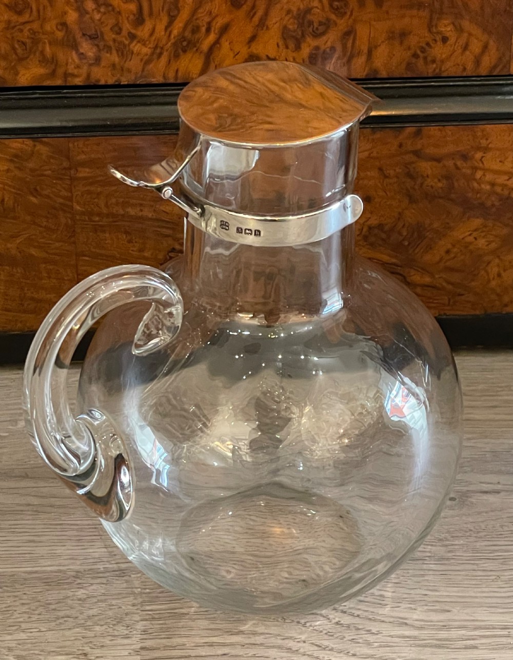 An Edwardian clear glass claret jug featuring a bulbous body, an integrated glass handle, and a polished silver-plated collar with a hinged spout lid. View V
