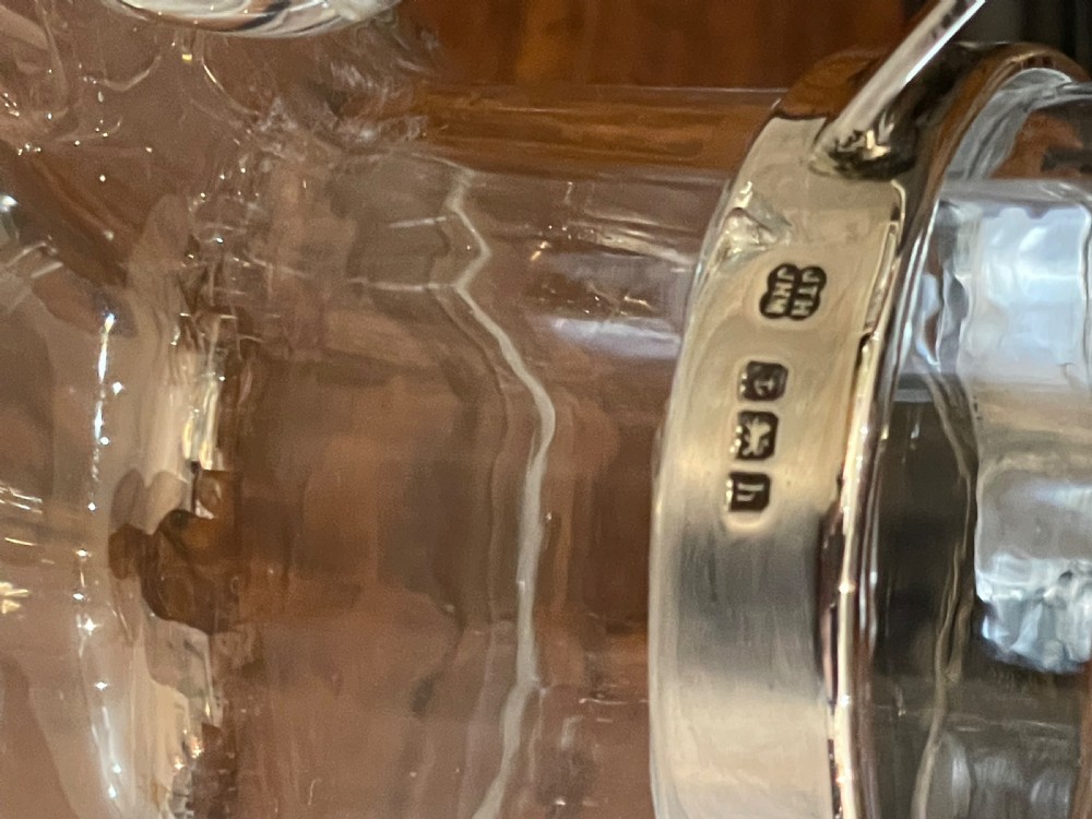 An Edwardian clear glass claret jug featuring a bulbous body, an integrated glass handle, and a polished silver-plated collar with a hinged spout lid. View VI