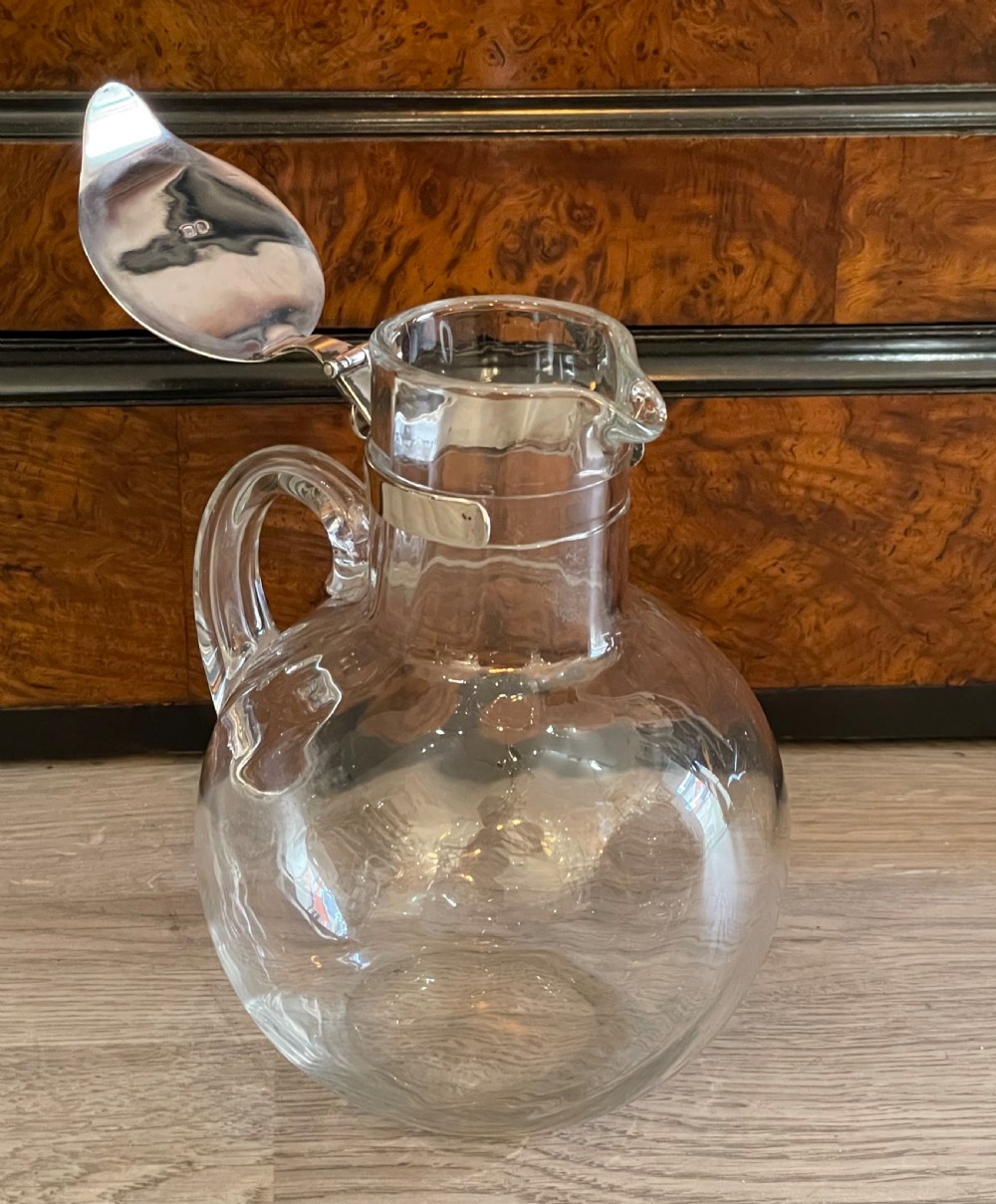 An Edwardian clear glass claret jug featuring a bulbous body, an integrated glass handle, and a polished silver-plated collar with a hinged spout lid. View VII