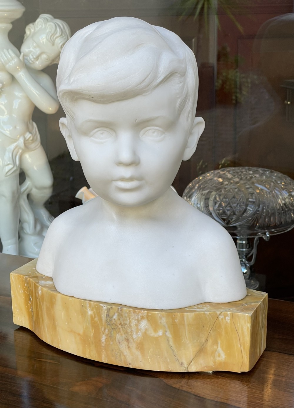 A white Carrara marble sculpture depicting the bust of a young boy, mounted on a yellow variegated marble base.