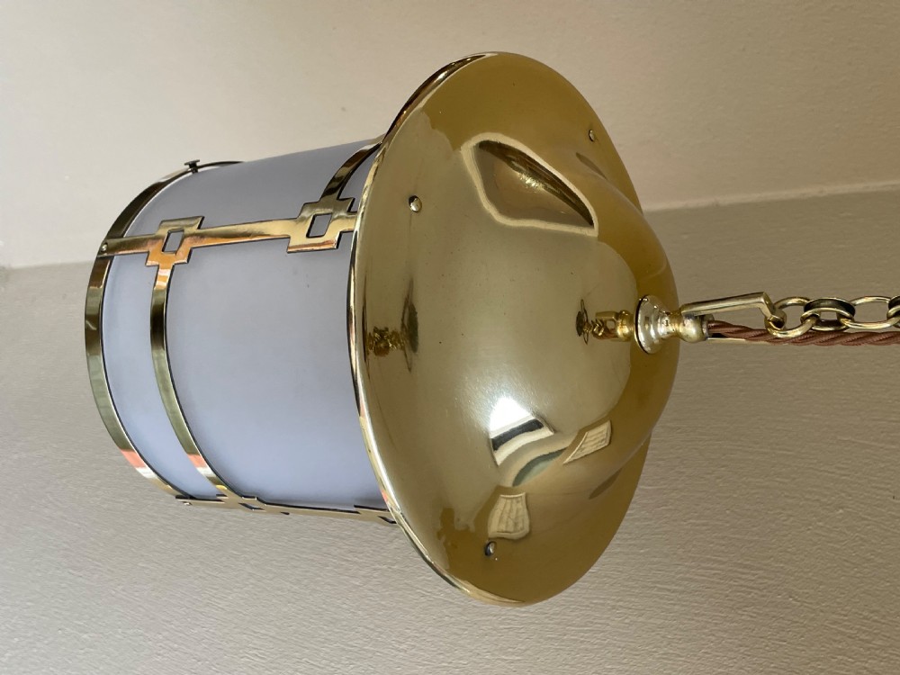 An Art Deco cylindrical hall lantern constructed with polished brass geometric framing surrounding a frosted glass shade, suspended by a brass chain. View V