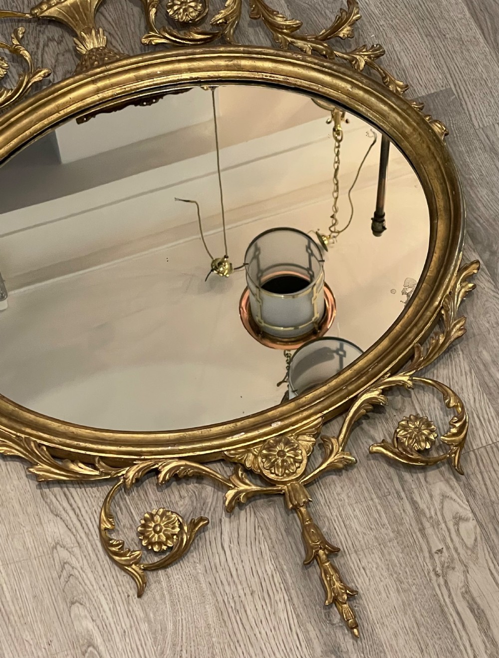 An oval giltwood wall mirror featuring an ornate Neoclassical frame topped with an urn and leaf motif and finished with a carved bottom finial. View II