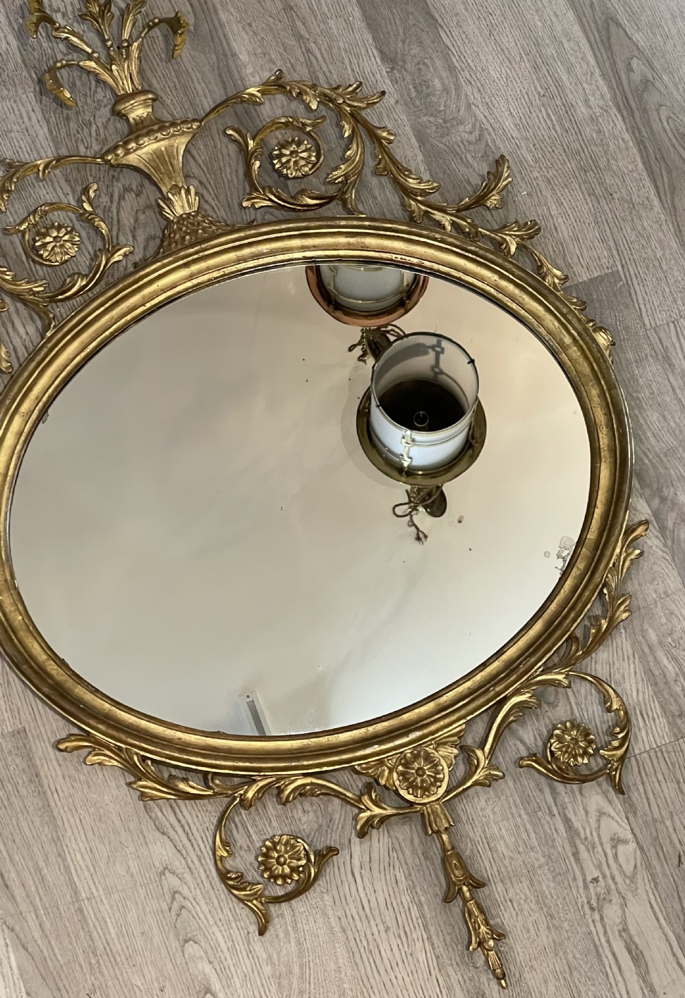 An oval giltwood wall mirror featuring an ornate Neoclassical frame topped with an urn and leaf motif and finished with a carved bottom finial. View III