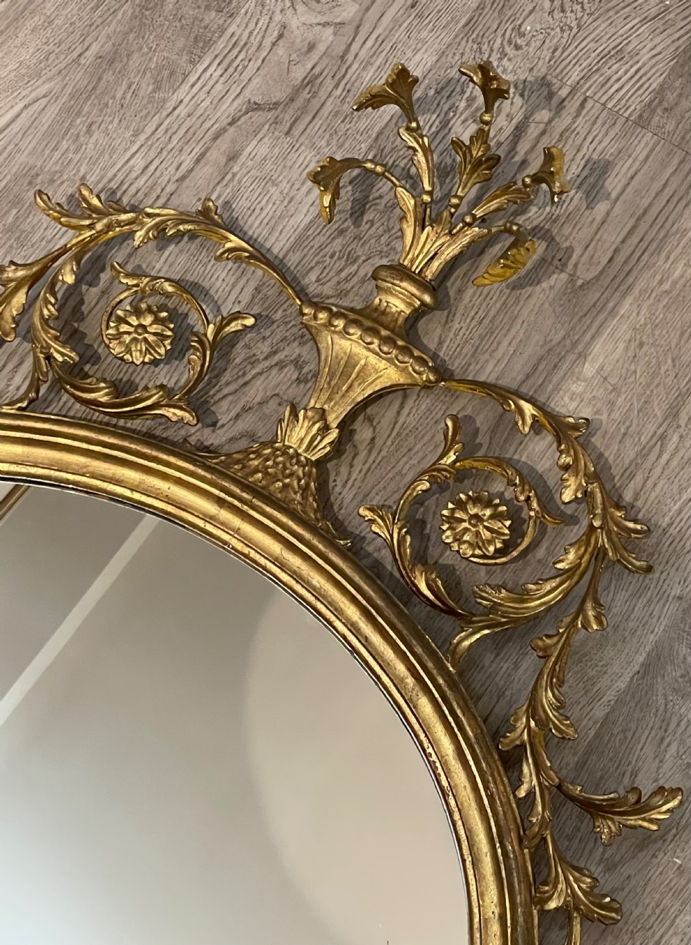 An oval giltwood wall mirror featuring an ornate Neoclassical frame topped with an urn and leaf motif and finished with a carved bottom finial. View IV