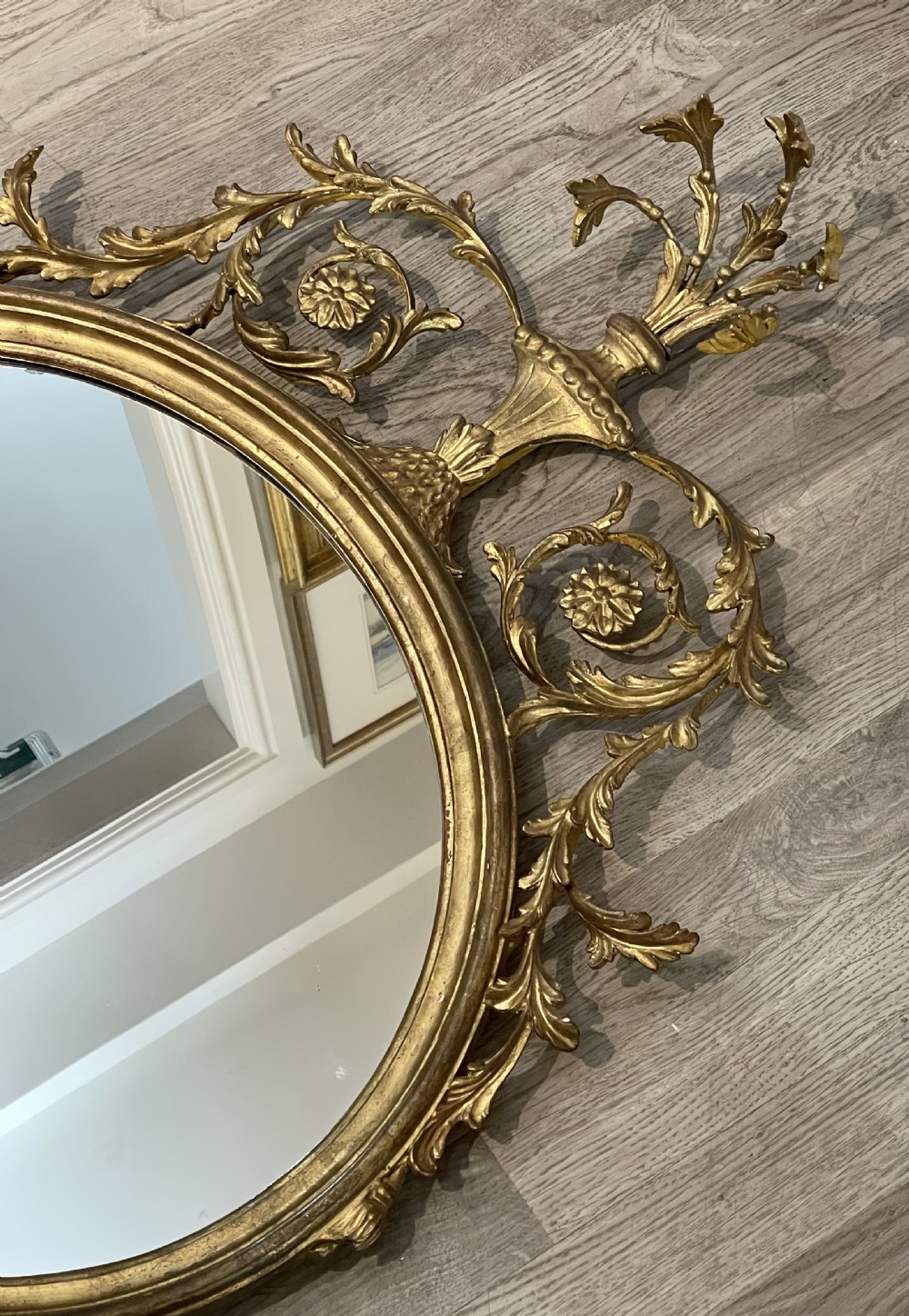 An oval giltwood wall mirror featuring an ornate Neoclassical frame topped with an urn and leaf motif and finished with a carved bottom finial. View V
