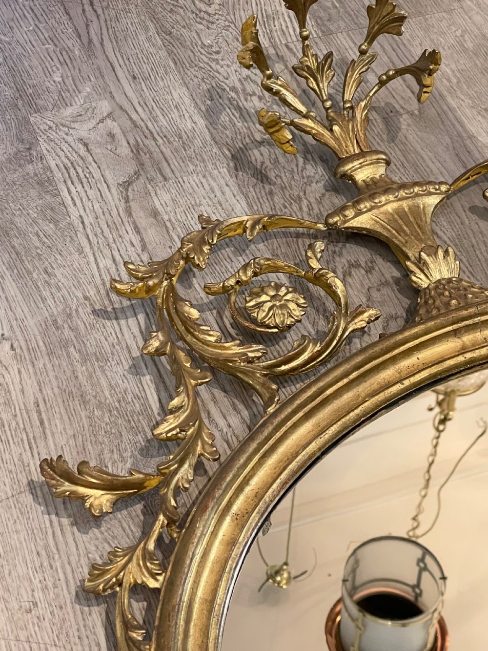 An oval giltwood wall mirror featuring an ornate Neoclassical frame topped with an urn and leaf motif and finished with a carved bottom finial. View VII