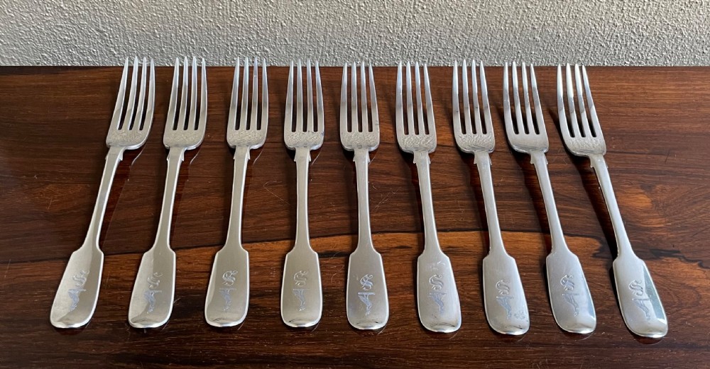 A set of nine antique Irish sterling silver dinner forks featuring fiddle pattern handles with engraved crests arranged on a dark wooden surface.