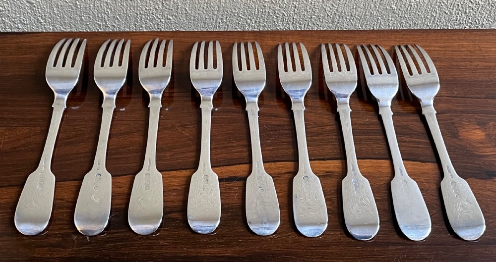 A set of nine antique Irish sterling silver dinner forks featuring fiddle pattern handles with engraved crests arranged on a dark wooden surface. View III