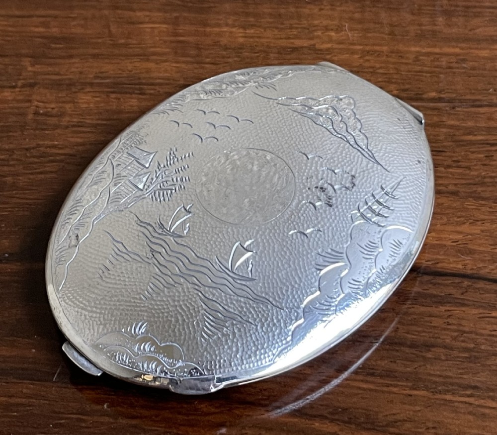 An oval silver compact featuring an engraved landscape scene with junk boats, mountains, trees, and a textured background.
