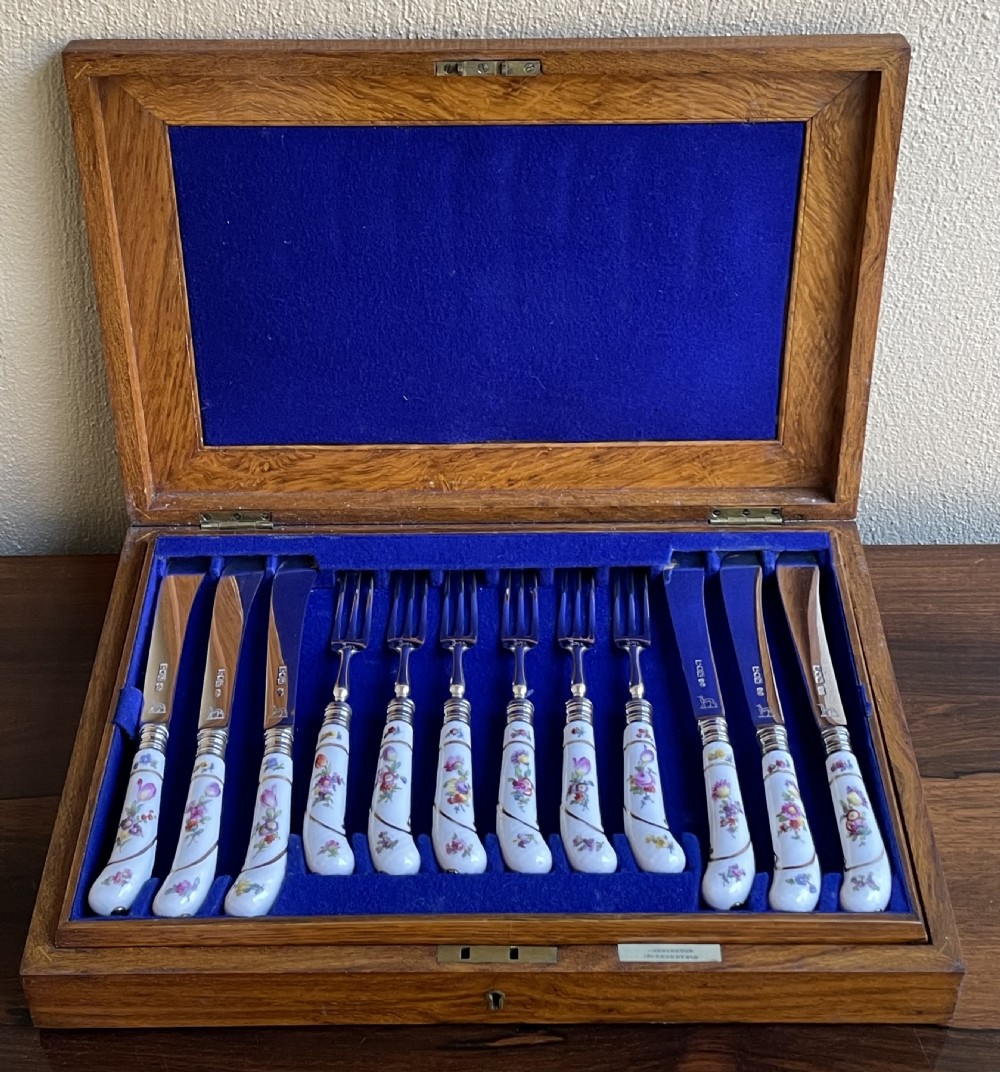 A twelve-piece silver and hand-painted porcelain dessert cutlery set housed in an open oak presentation box lined with blue velvet.