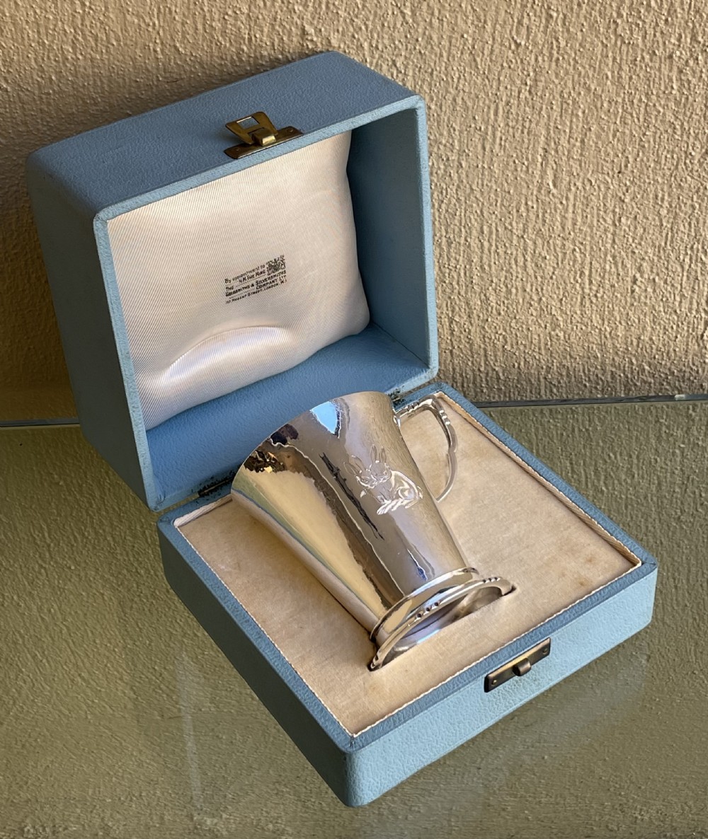 A tapered sterling silver christening mug with an engraved rabbit motif resting inside a light blue presentation case.