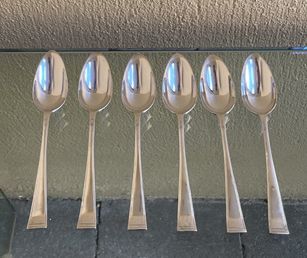 A set of six silver Art Deco grapefruit spoons with geometric tapered handles arranged in a row on a reflective surface.