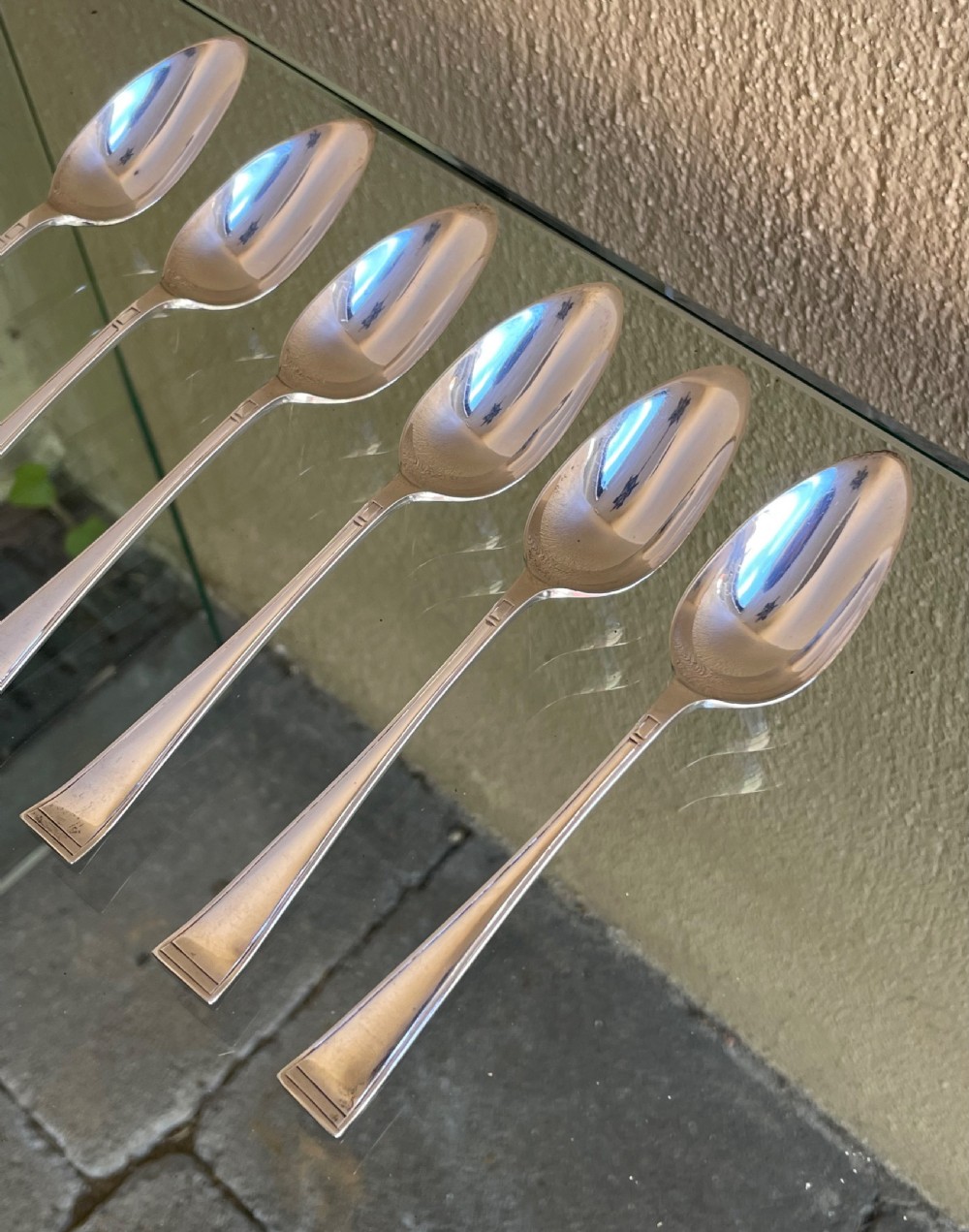A set of six silver Art Deco grapefruit spoons with geometric tapered handles arranged in a row on a reflective surface. View II