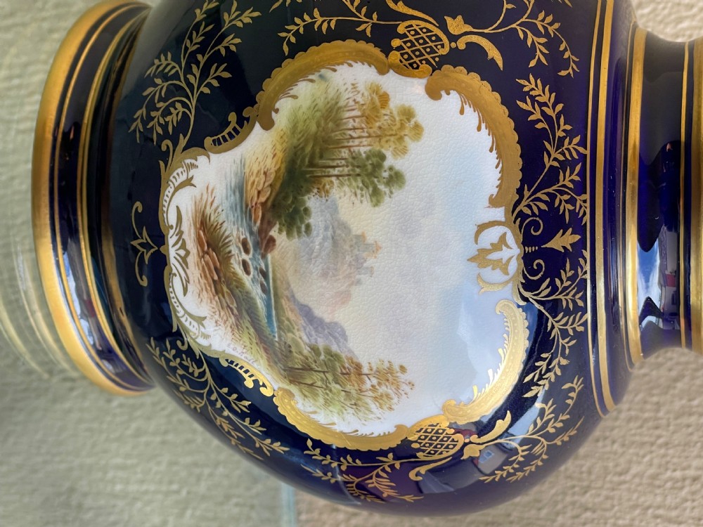 A bulbous cobalt blue porcelain vase with a fitted lid, featuring ornate gilded foliate borders surrounding a central hand-painted landscape cartouche. View II