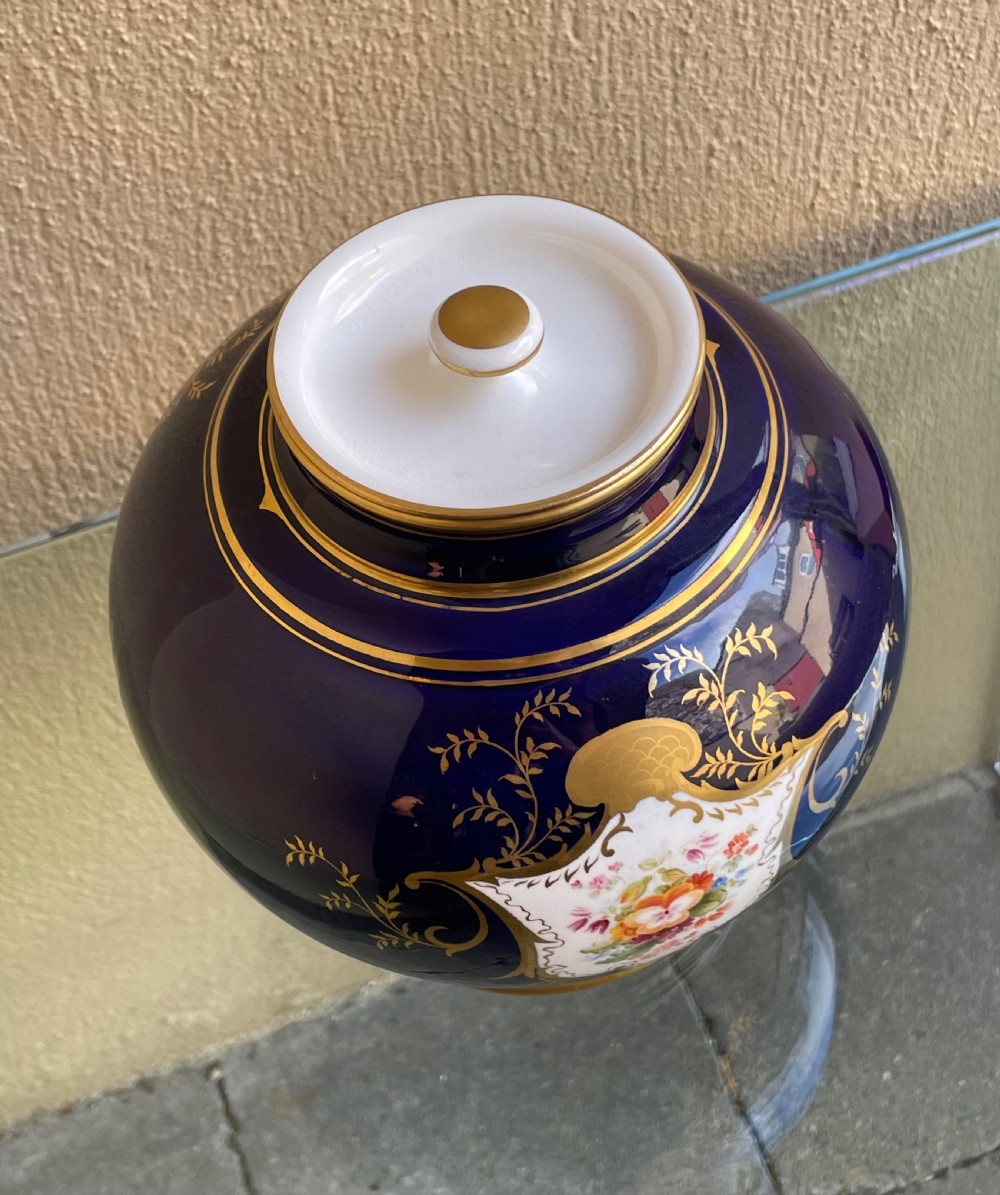 A bulbous cobalt blue porcelain vase with a fitted lid, featuring ornate gilded foliate borders surrounding a central hand-painted landscape cartouche. View IV