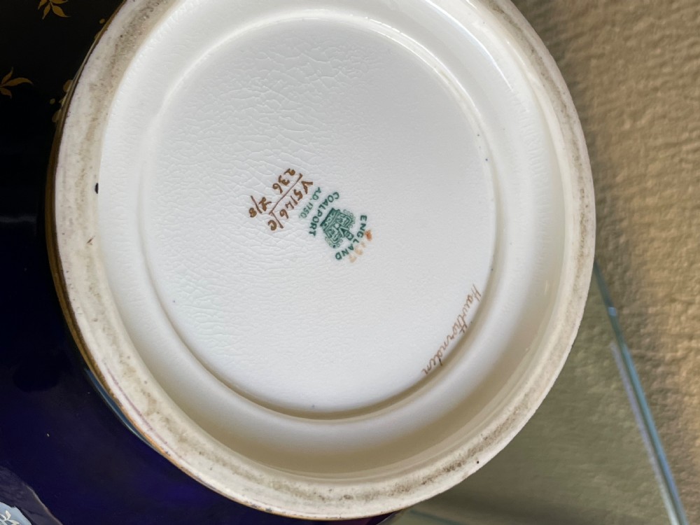 A bulbous cobalt blue porcelain vase with a fitted lid, featuring ornate gilded foliate borders surrounding a central hand-painted landscape cartouche. View VI