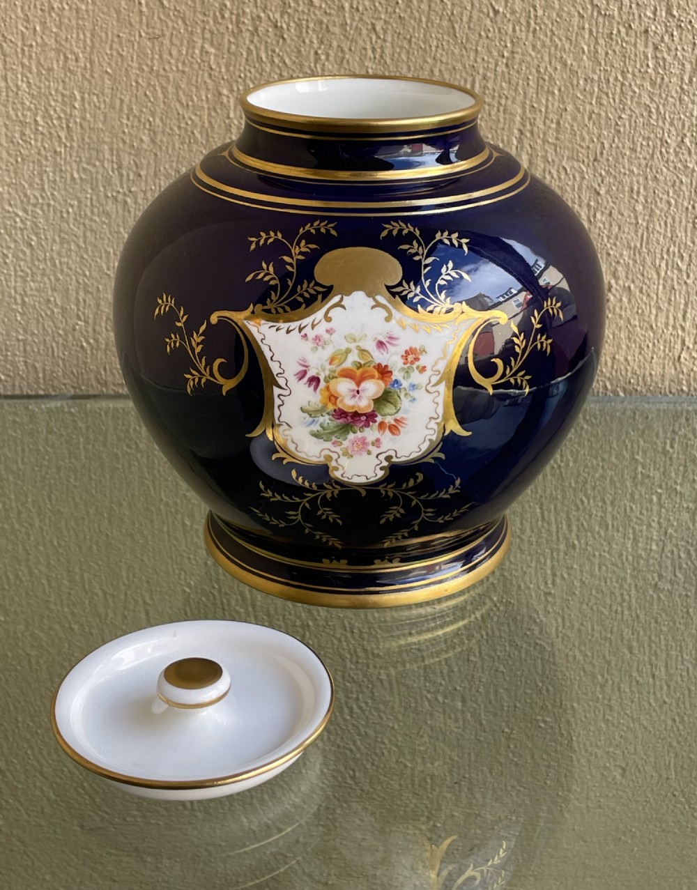 A bulbous cobalt blue porcelain vase with a fitted lid, featuring ornate gilded foliate borders surrounding a central hand-painted landscape cartouche. View VII