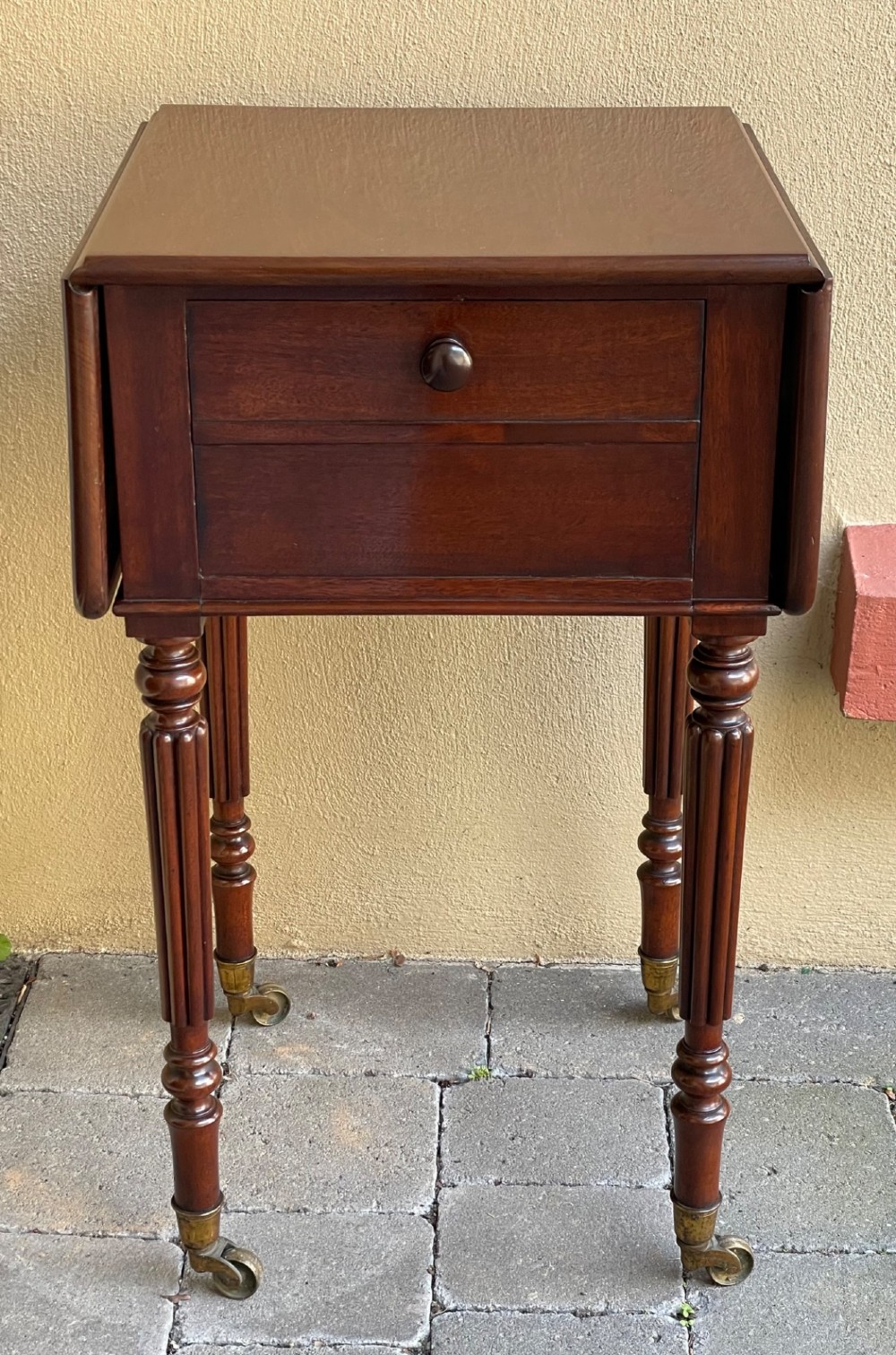 An early Victorian mahogany bedside table featuring drop-leaf sides, a single drawer with a wooden knob, reeded turned legs, and brass castors.