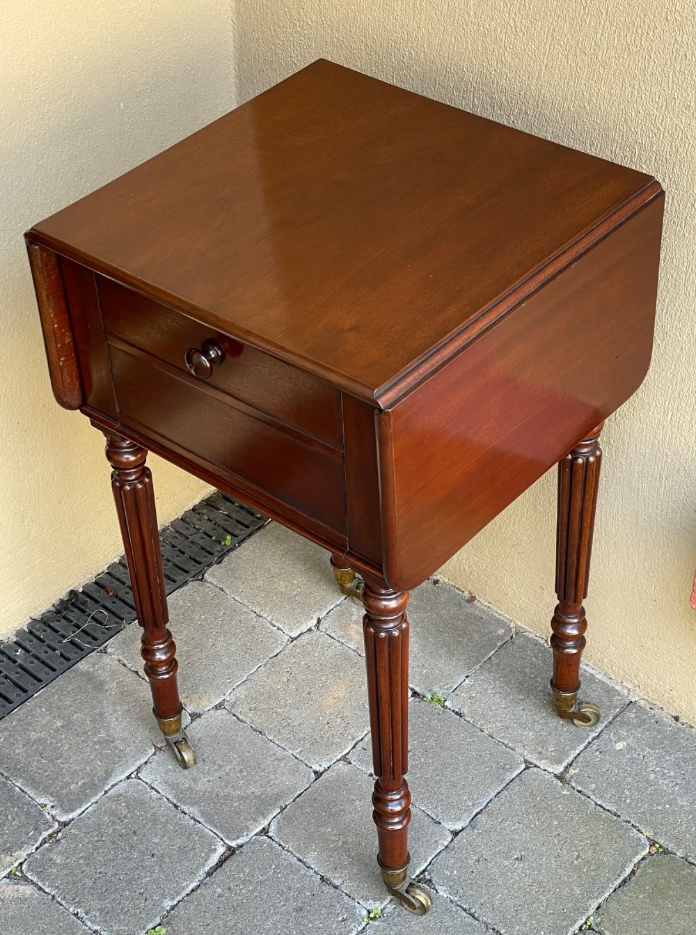An early Victorian mahogany bedside table featuring drop-leaf sides, a single drawer with a wooden knob, reeded turned legs, and brass castors. View II