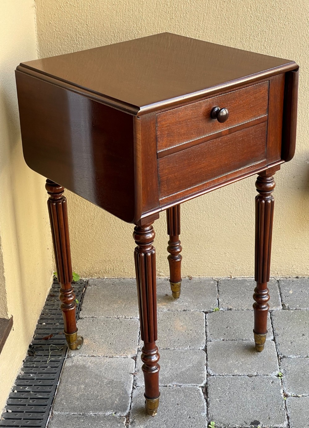 An early Victorian mahogany bedside table featuring drop-leaf sides, a single drawer with a wooden knob, reeded turned legs, and brass castors. View III