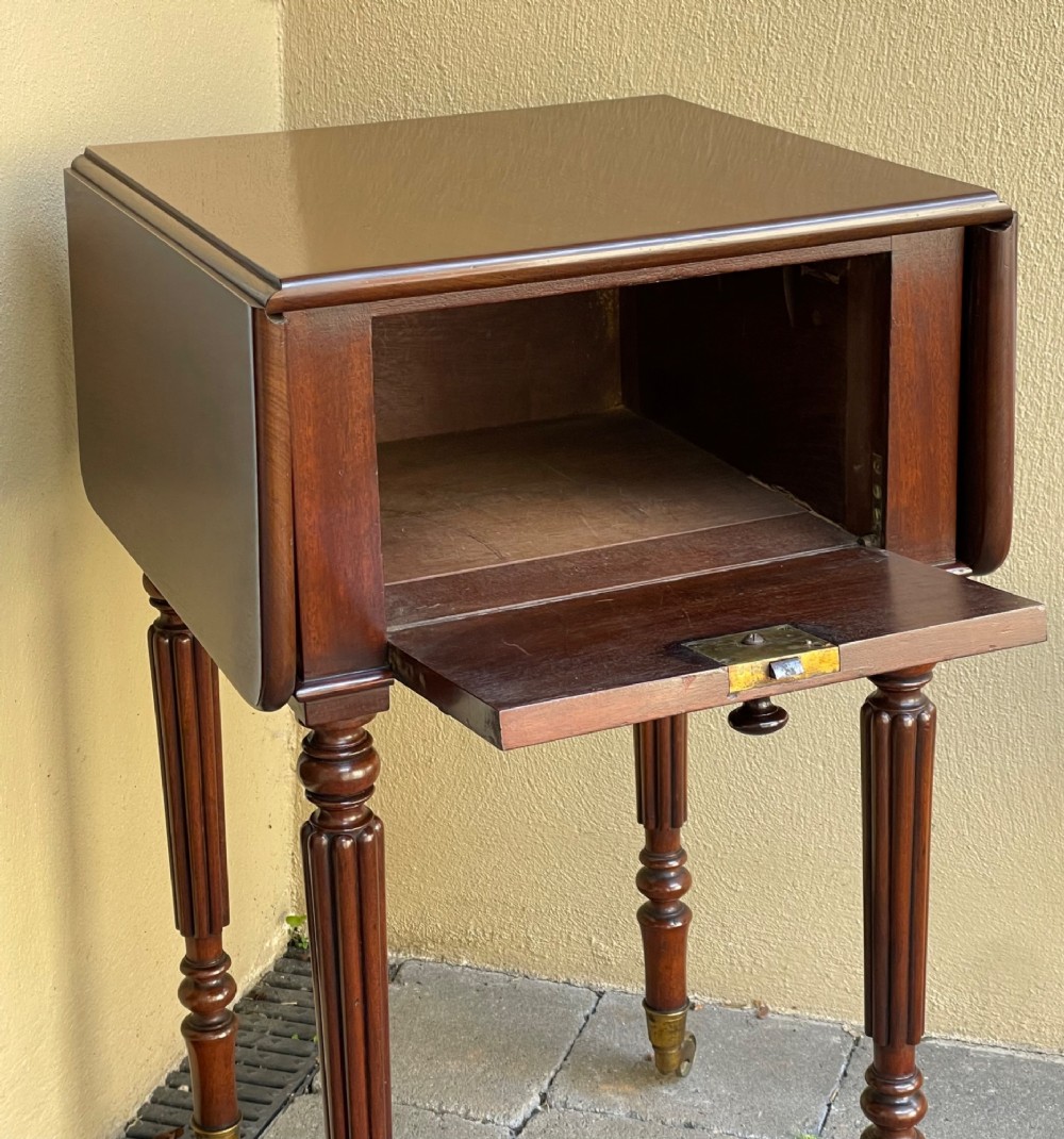 An early Victorian mahogany bedside table featuring drop-leaf sides, a single drawer with a wooden knob, reeded turned legs, and brass castors. View IV