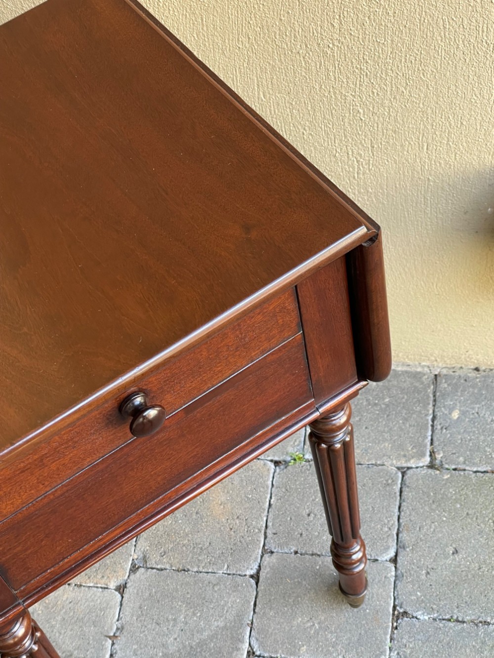 An early Victorian mahogany bedside table featuring drop-leaf sides, a single drawer with a wooden knob, reeded turned legs, and brass castors. View V