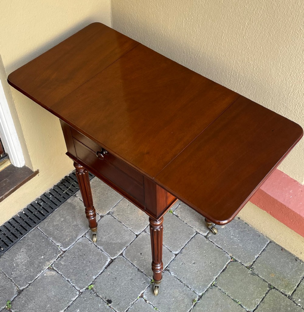An early Victorian mahogany bedside table featuring drop-leaf sides, a single drawer with a wooden knob, reeded turned legs, and brass castors. View VI