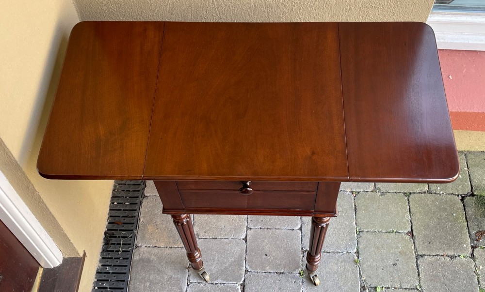 An early Victorian mahogany bedside table featuring drop-leaf sides, a single drawer with a wooden knob, reeded turned legs, and brass castors. View VII