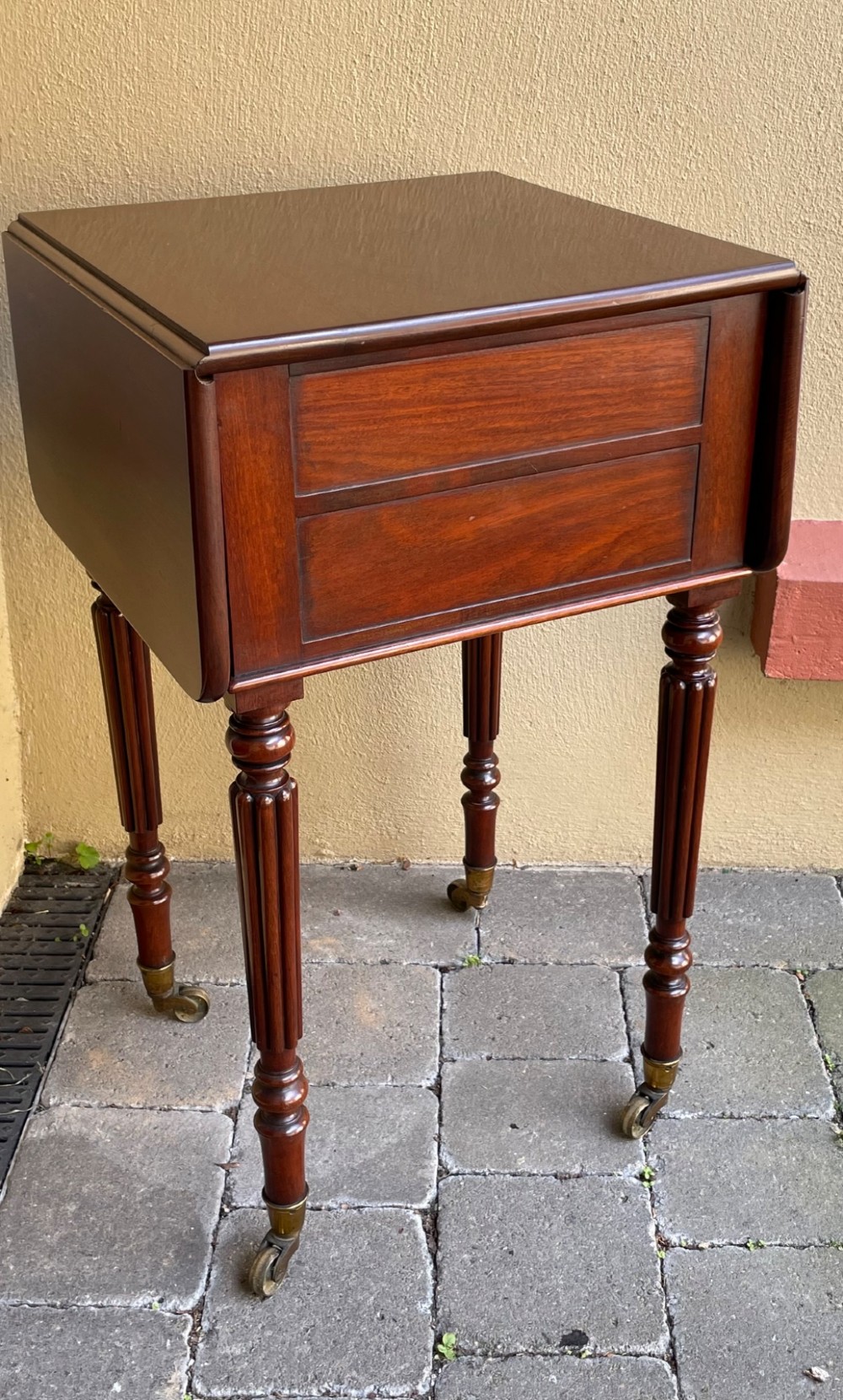 An early Victorian mahogany bedside table featuring drop-leaf sides, a single drawer with a wooden knob, reeded turned legs, and brass castors. View VIII