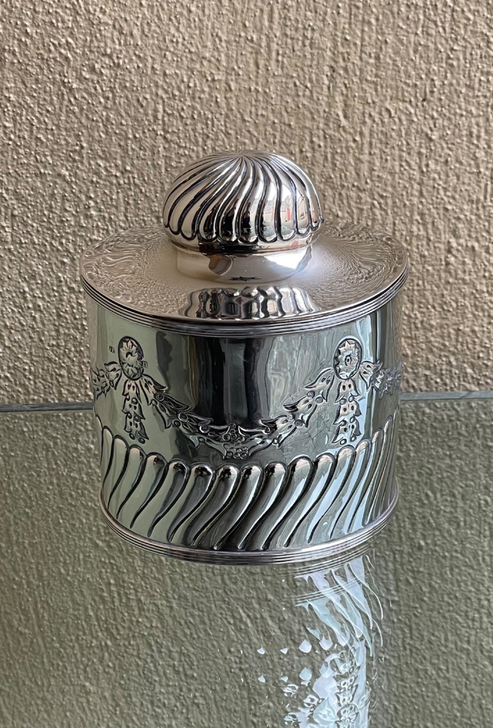 A cylindrical Edwardian sterling silver tea caddy featuring embossed floral garlands and spiral fluting around the lower body.