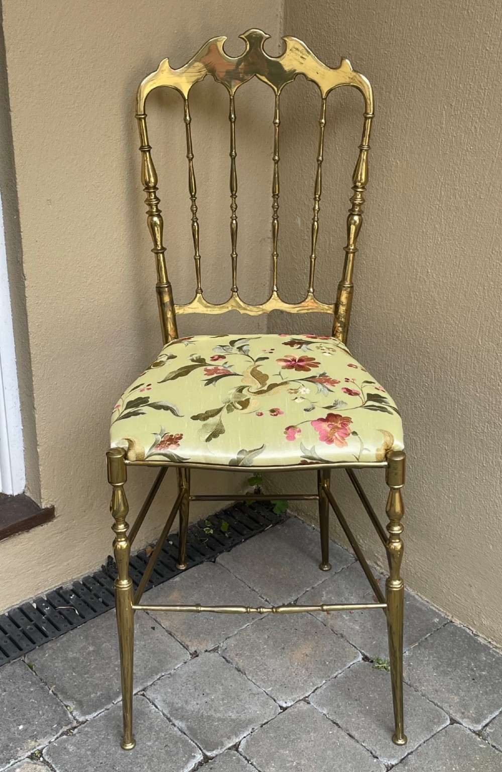 A polished brass Chiavari style salon chair featuring a spindle backrest, ornate cresting rail, turned legs, and a floral upholstered seat.