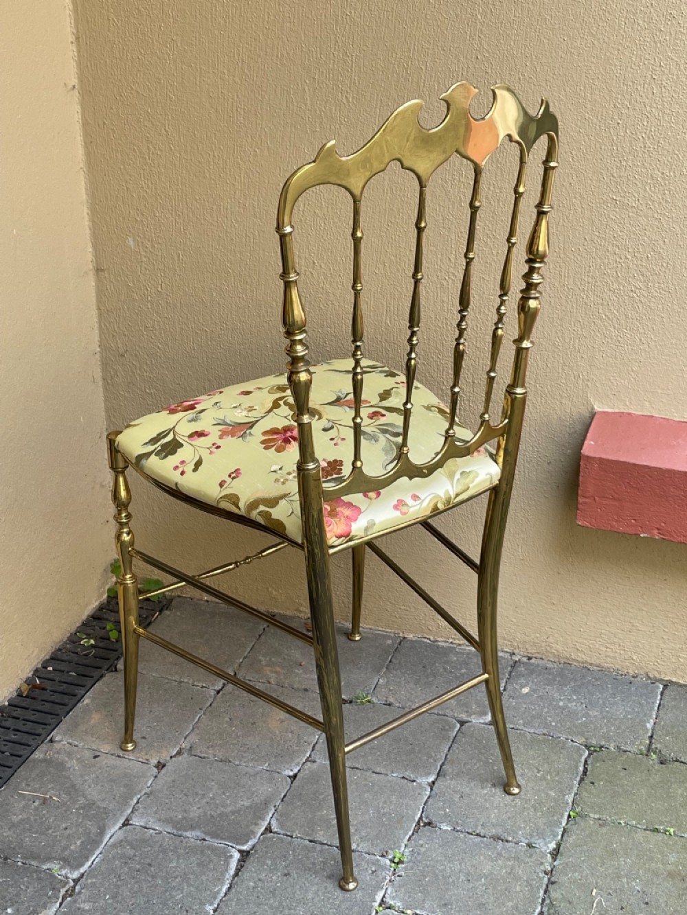 A polished brass Chiavari style salon chair featuring a spindle backrest, ornate cresting rail, turned legs, and a floral upholstered seat. View VI