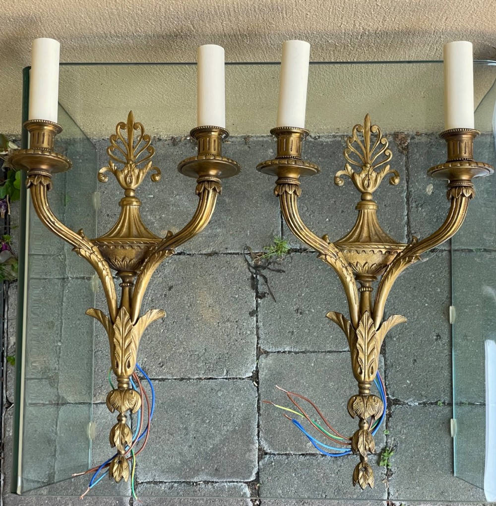 A pair of French Neoclassical gilded bronze wall sconces featuring urn-shaped central bodies, twin branches, acanthus leaf embellishments, and white candle sleeves.