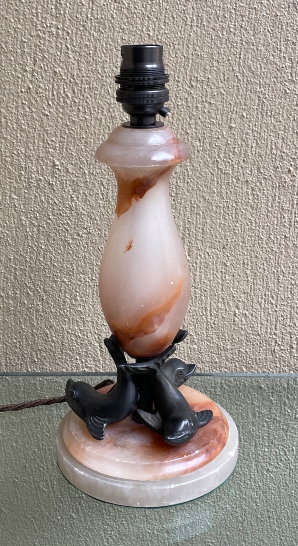 An Art Deco table lamp featuring a veined pink and cream alabaster vase column resting on three dark patinated metal dolphins over a round alabaster base.