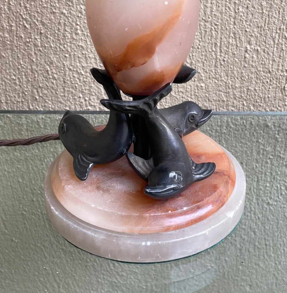 An Art Deco table lamp featuring a veined pink and cream alabaster vase column resting on three dark patinated metal dolphins over a round alabaster base. View III