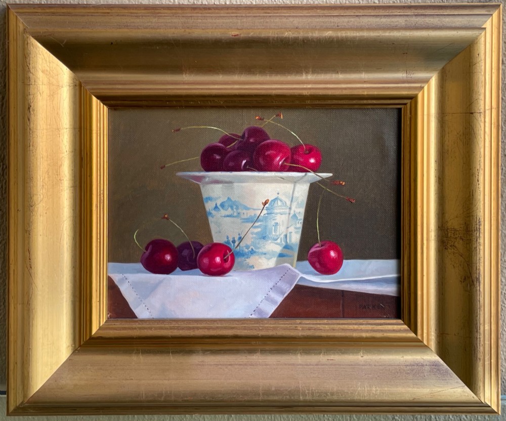 A framed oil painting featuring a composition of ripe red cherries in and around a blue and white patterned ceramic cup, presented in a broad gold gilt frame.