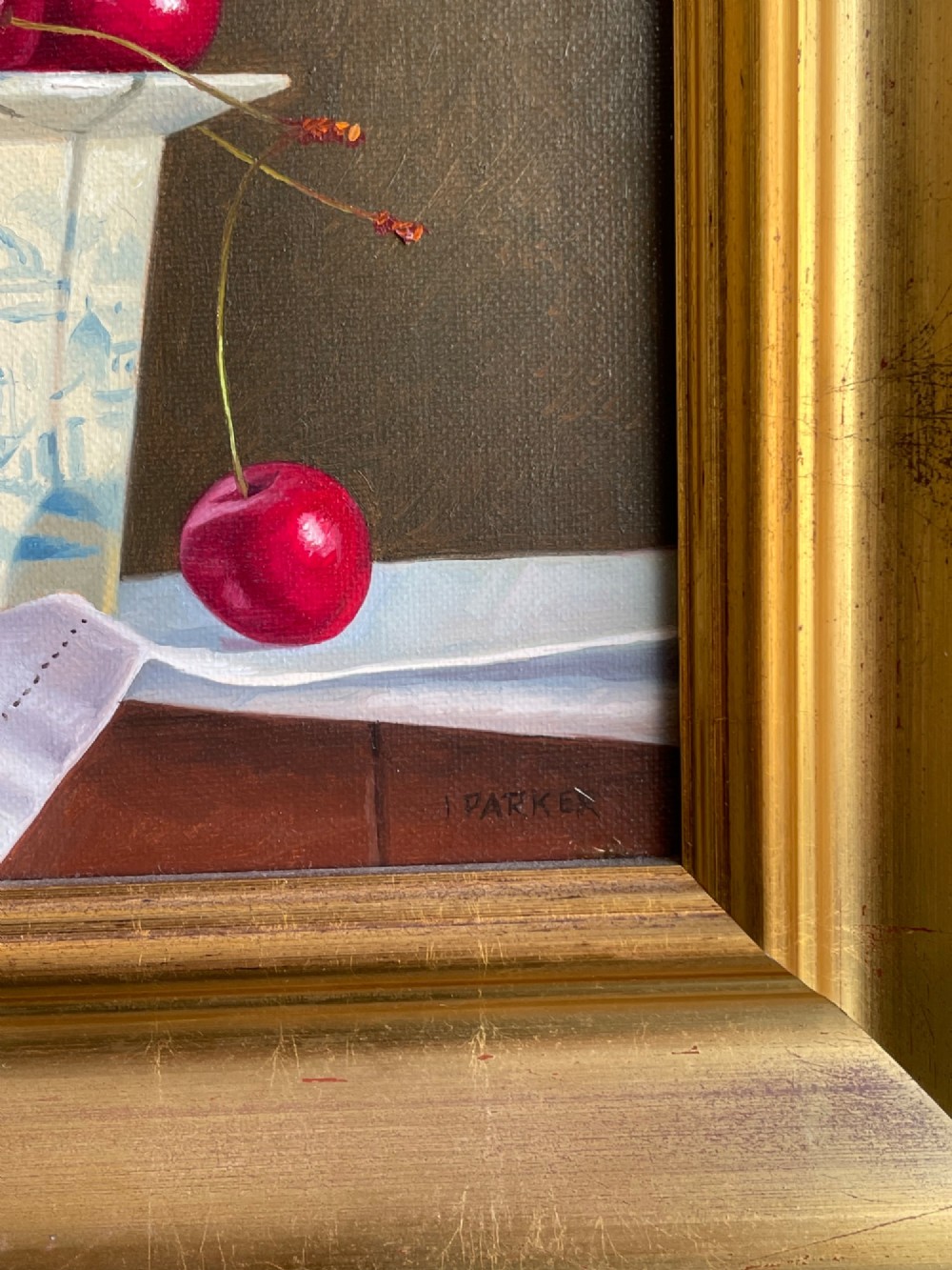 A framed oil painting featuring a composition of ripe red cherries in and around a blue and white patterned ceramic cup, presented in a broad gold gilt frame. View II