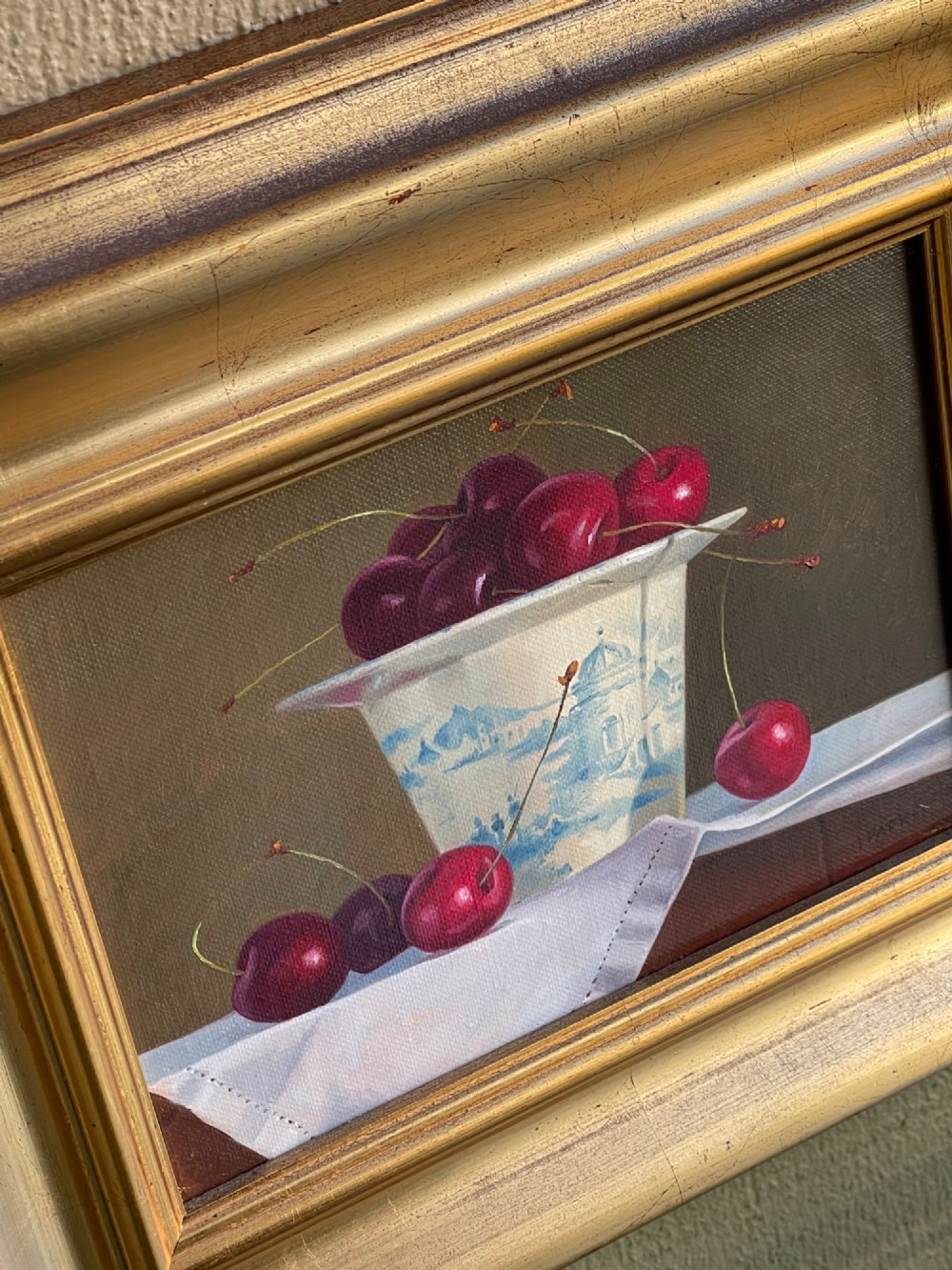 A framed oil painting featuring a composition of ripe red cherries in and around a blue and white patterned ceramic cup, presented in a broad gold gilt frame. View III