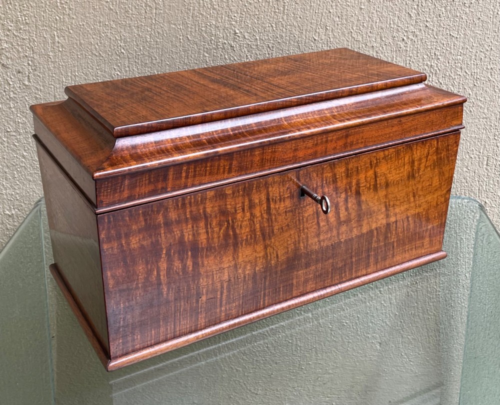 A rectangular Regency flame mahogany tea caddy featuring a stepped hinged lid and a brass key inserted into the front lock.