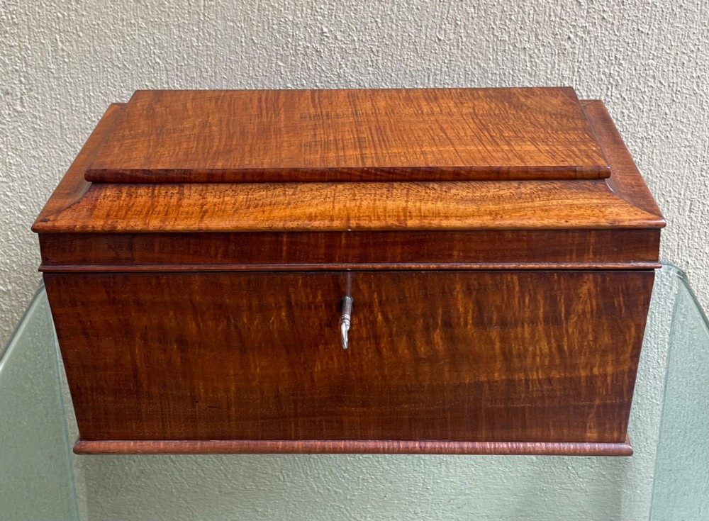 A rectangular Regency flame mahogany tea caddy featuring a stepped hinged lid and a brass key inserted into the front lock. View II