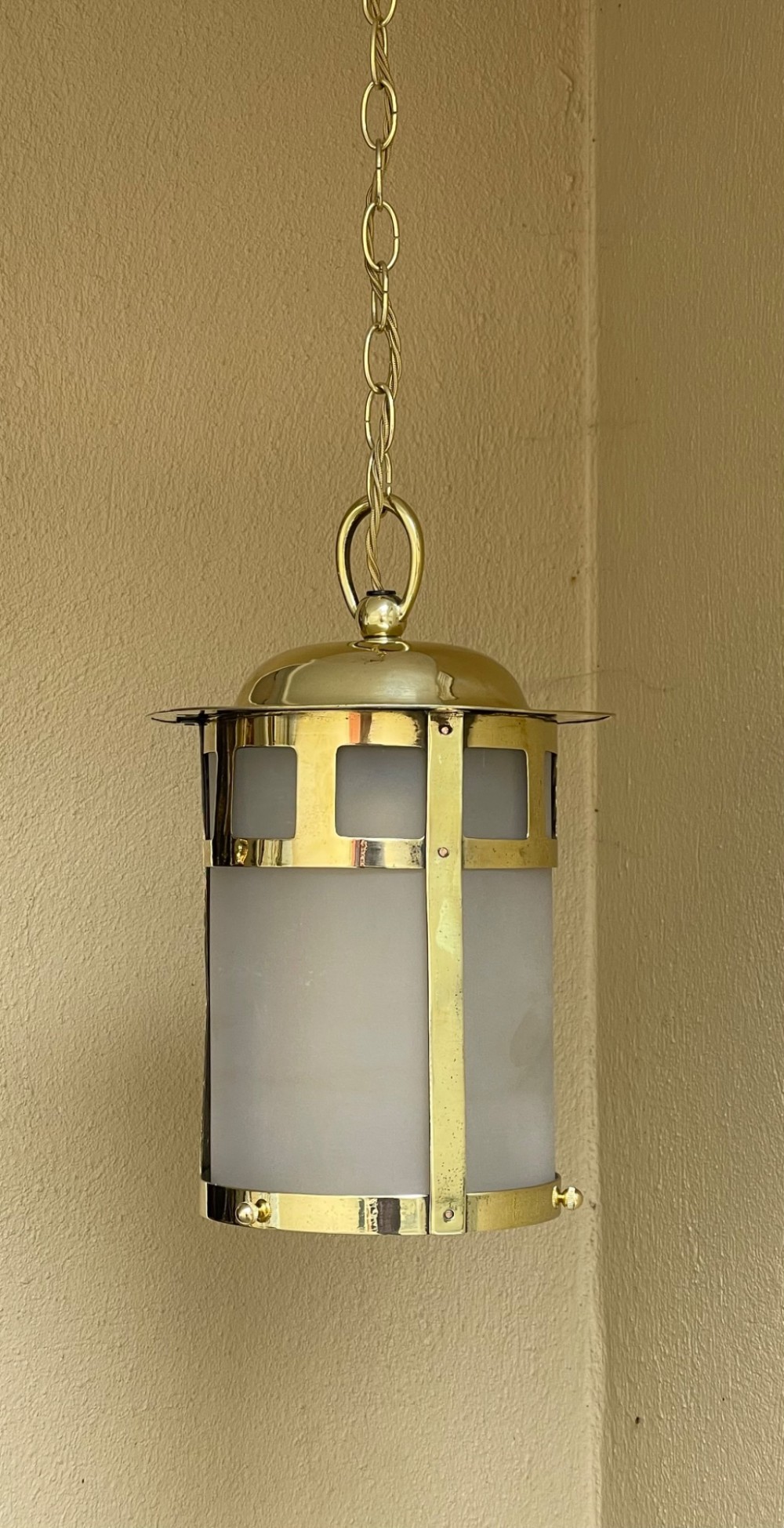 An Arts and Crafts style brass hall lantern with geometric cut-out metalwork and a frosted glass cylindrical shade, suspended by a brass chain. View III