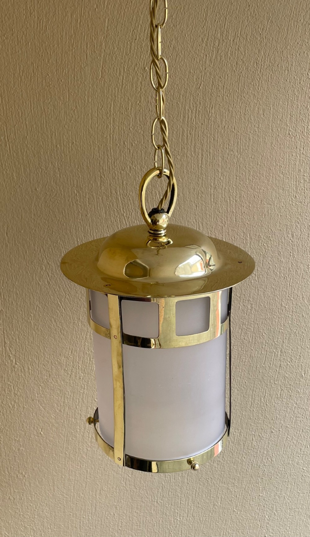 An Arts and Crafts style brass hall lantern with geometric cut-out metalwork and a frosted glass cylindrical shade, suspended by a brass chain. View IV