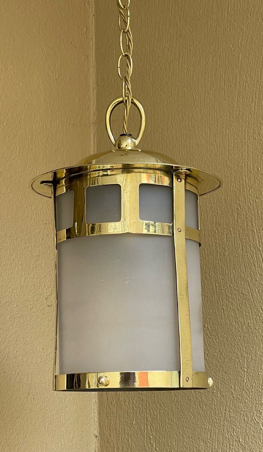 An Arts and Crafts style brass hall lantern with geometric cut-out metalwork and a frosted glass cylindrical shade, suspended by a brass chain. View V