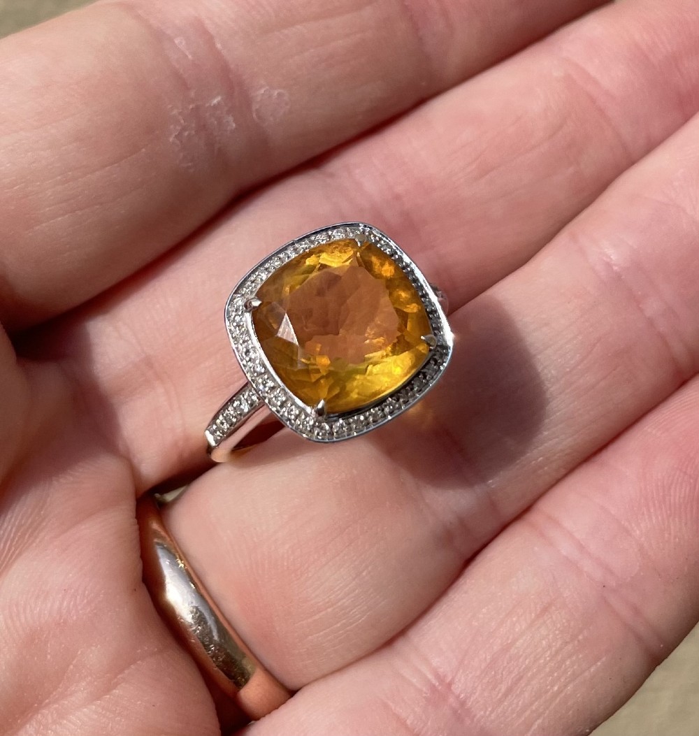 A cushion-cut yellow citrine gemstone set in a white metal ring surrounded by a border of small brilliant-cut diamonds.