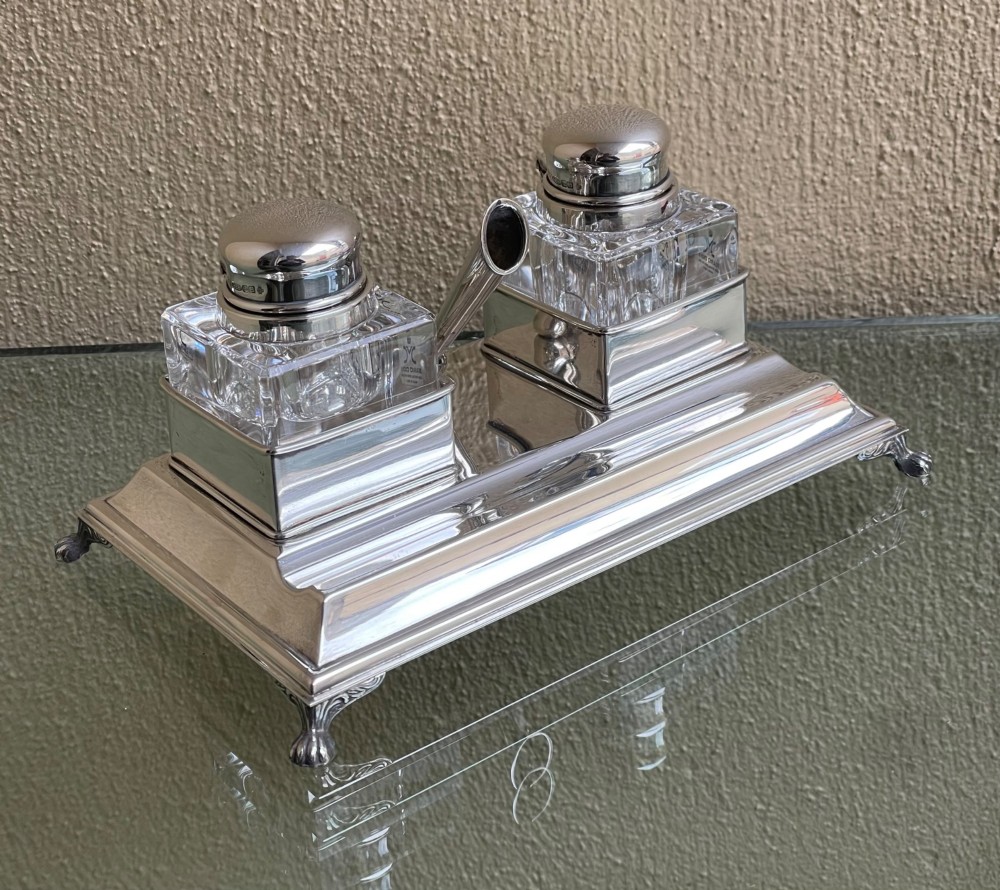 A hallmarked solid silver inkstand featuring two square cut-glass inkwells with silver lids mounted on a stepped tray with decorative feet.